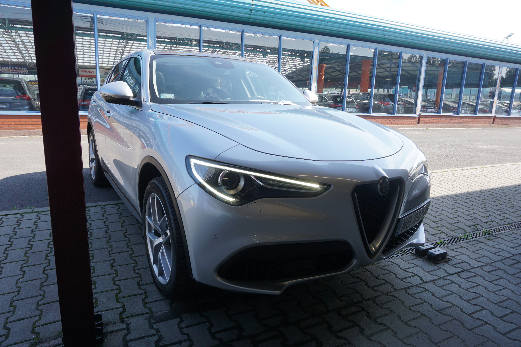 Srebrny SUV Alfa Romeo Stelvio zaparkowany przed salonem samochodowym z przeszkloną fasadą, widok z przodu, oświetlenie dzienne.