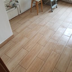 Marek Baczkiewicz - Ułożona podłoga z płytek imitujących drewno, widoczny fragment ściany z rurami, drabina malarska i wiadro z farbą na taborecie.