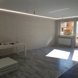 Marek Baczkiewicz - Wnętrze pokoju po odświeżeniu, z białą ceglaną ścianą, podwieszanym sufitem z oświetleniem LED, szarą podłogą imitującą deski z nadrukiem, widoczne przygotowane akcesoria na stole i wiszące żarówki...