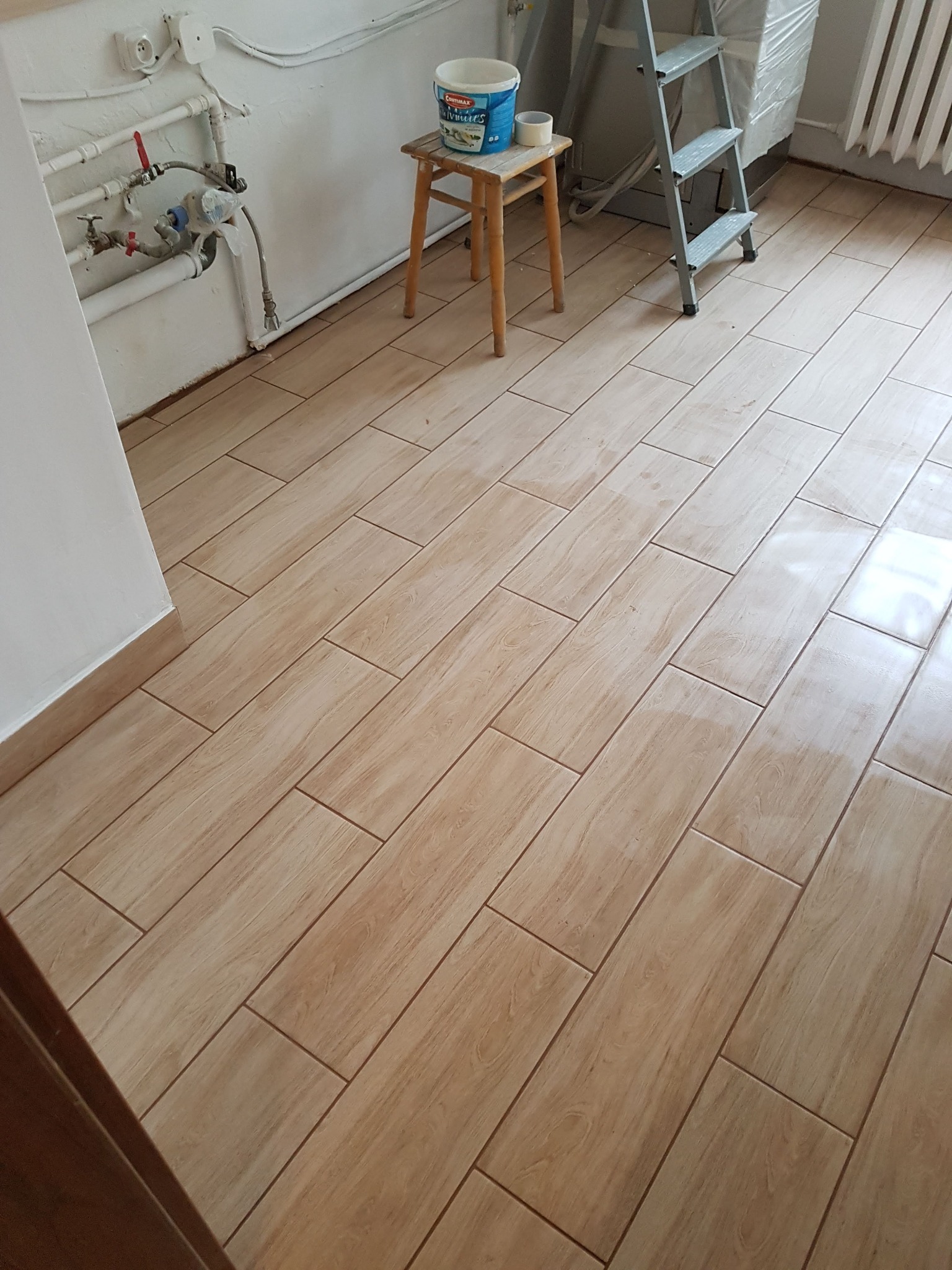 Ułożona podłoga z płytek imitujących drewno, widoczny fragment ściany z rurami, drabina malarska i wiadro z farbą na taborecie.