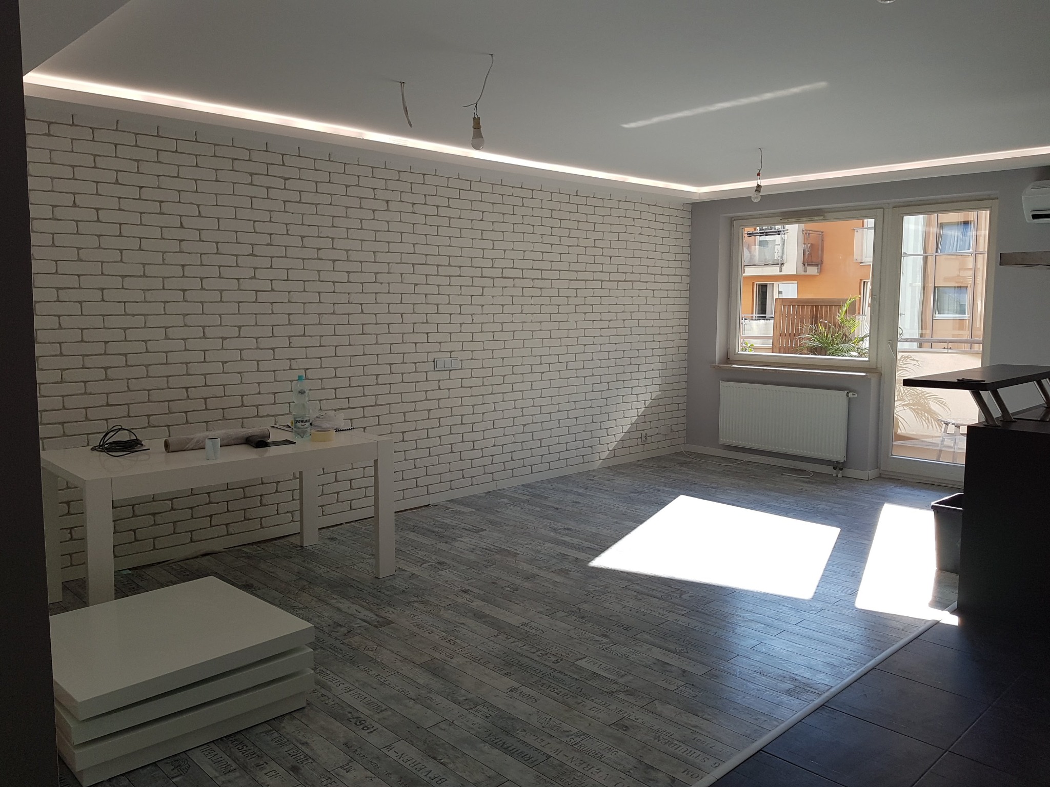 Wnętrze pokoju po odświeżeniu, z białą ceglaną ścianą, podwieszanym sufitem z oświetleniem LED, szarą podłogą imitującą deski z nadrukiem, widoczne przygotowane akcesoria na stole i wiszące żarówki...