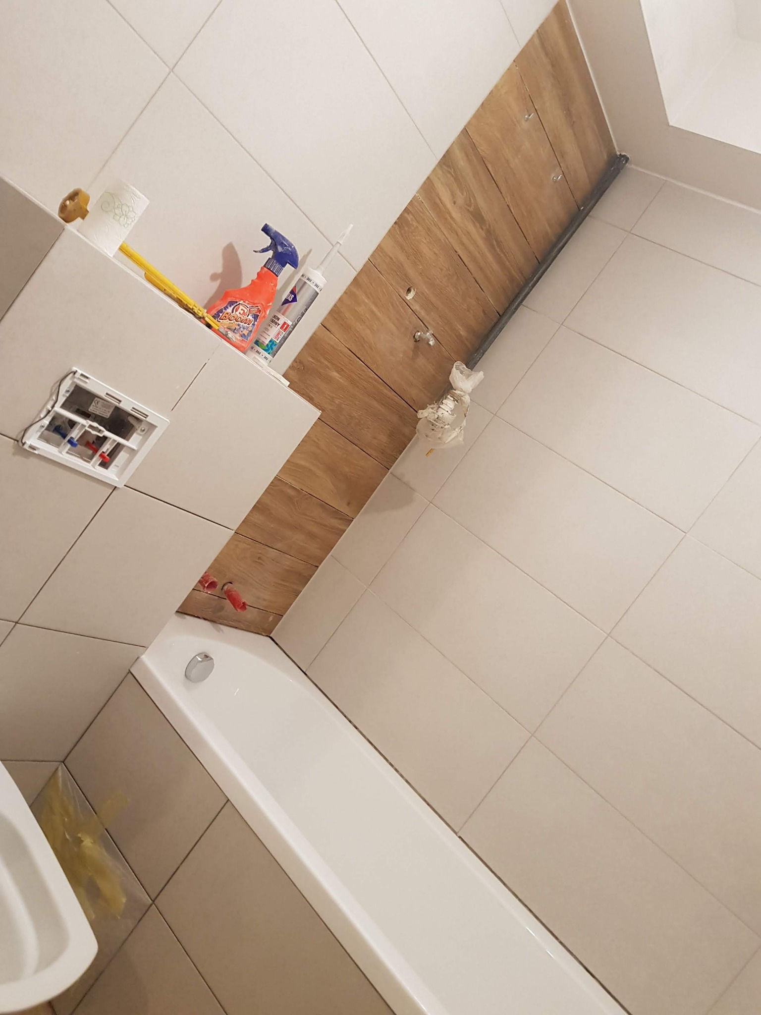 Montaż drewnopodobnych paneli na ścianie w łazience, widoczne narzędzia i chemia budowlana, biała wanna i umywalka, instalacja podtynkowa WC w trakcie montażu.