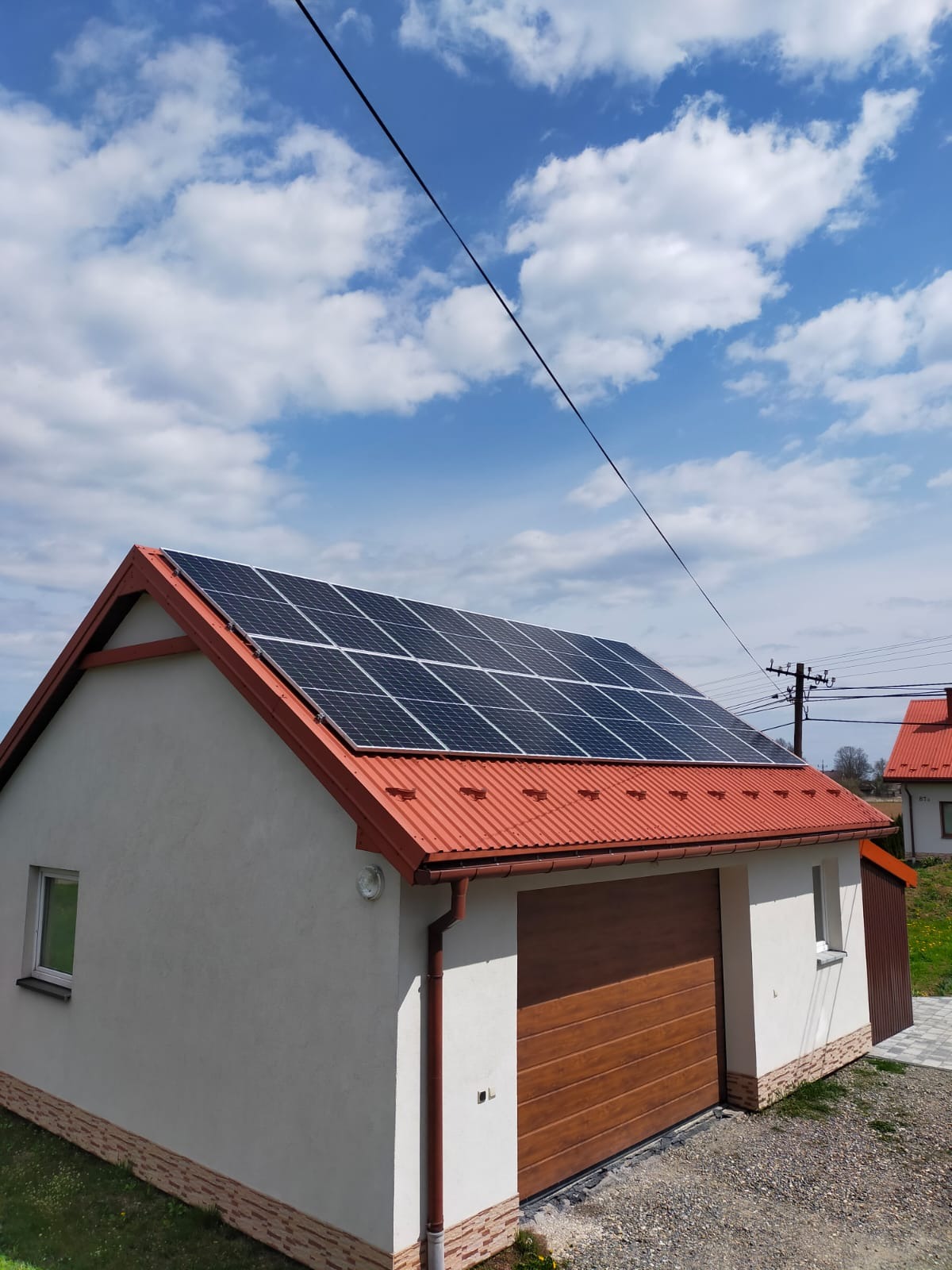 Panele słoneczne zamontowane na dachu garażu z czerwoną blachodachówką, widoczne błękitne niebo z chmurami.