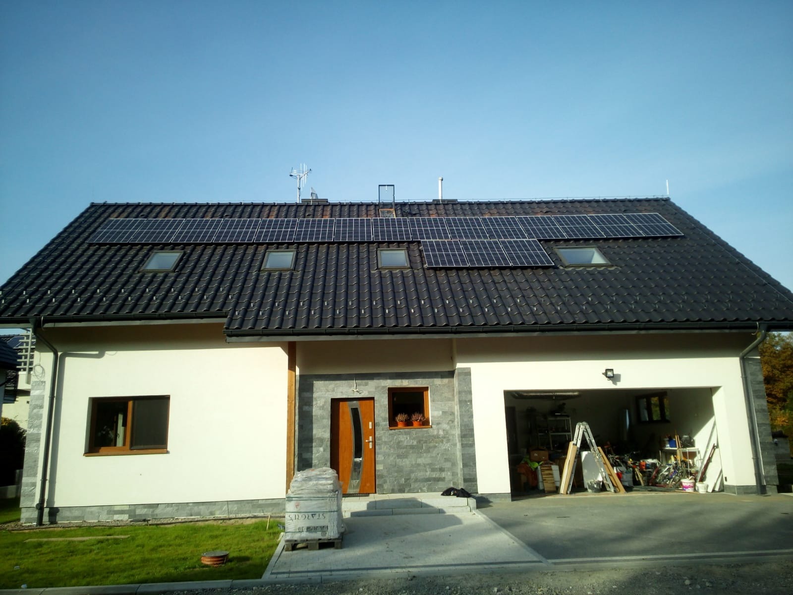 Dom jednorodzinny z ciemną dachówką, na której zamontowane są panele słoneczne, widoczne również okna dachowe i antena telewizyjna. W tle błękitne niebo.