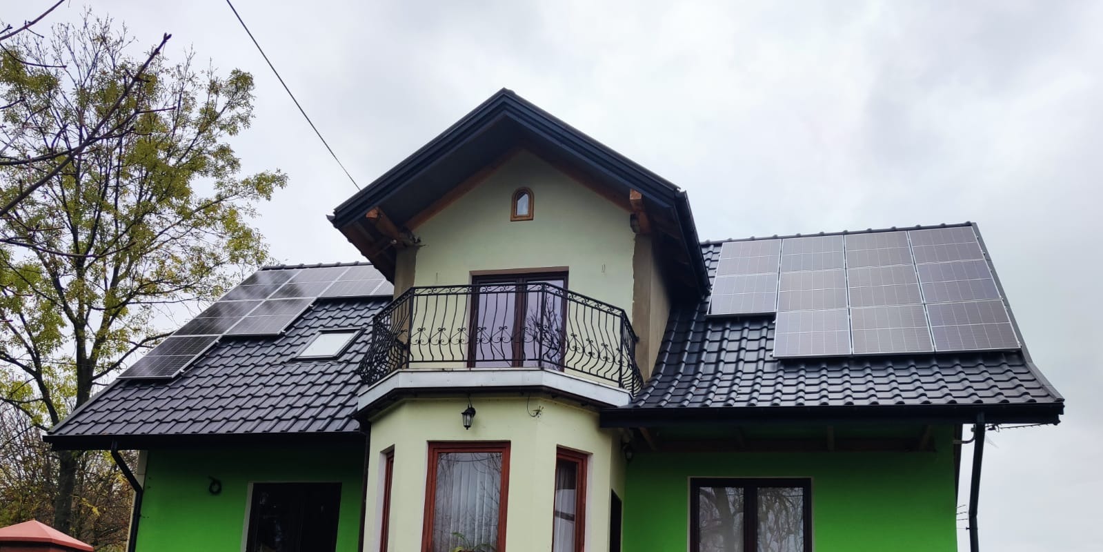 Dwukondygnacyjny dom z zieloną elewacją i panelami słonecznymi na dachu pokrytym czarną dachówką, widok na balkon z ozdobną metalową balustradą.