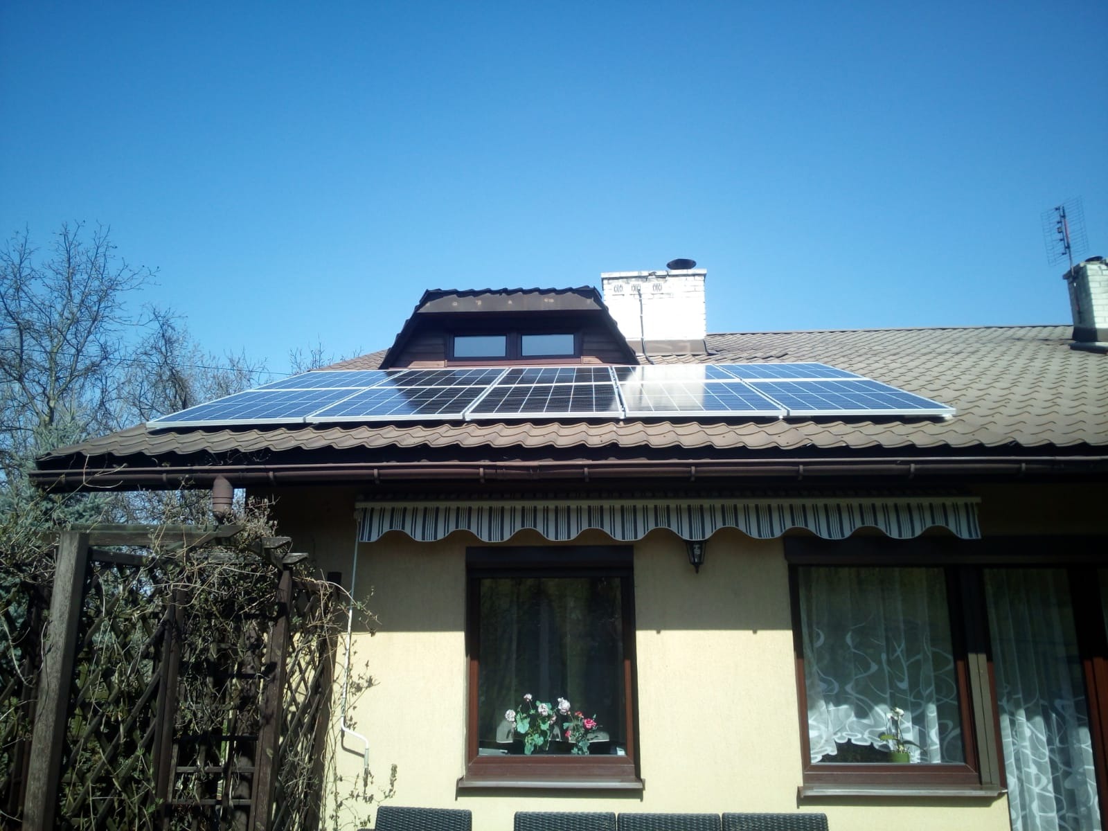 Panele fotowoltaiczne na brązowym dachu domu jednorodzinnego z oknem dachowym i kominem, widoczne rośliny w oknach.