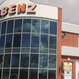 Budynek agencji celnej i ubezpieczeniowej Olbenz z dużymi oknami, w których odbija się błękitne niebo z chmurami. Na ścianie tabliczki informacyjne dotyczące agencji celnej J.K Cargo i Warta HDI...