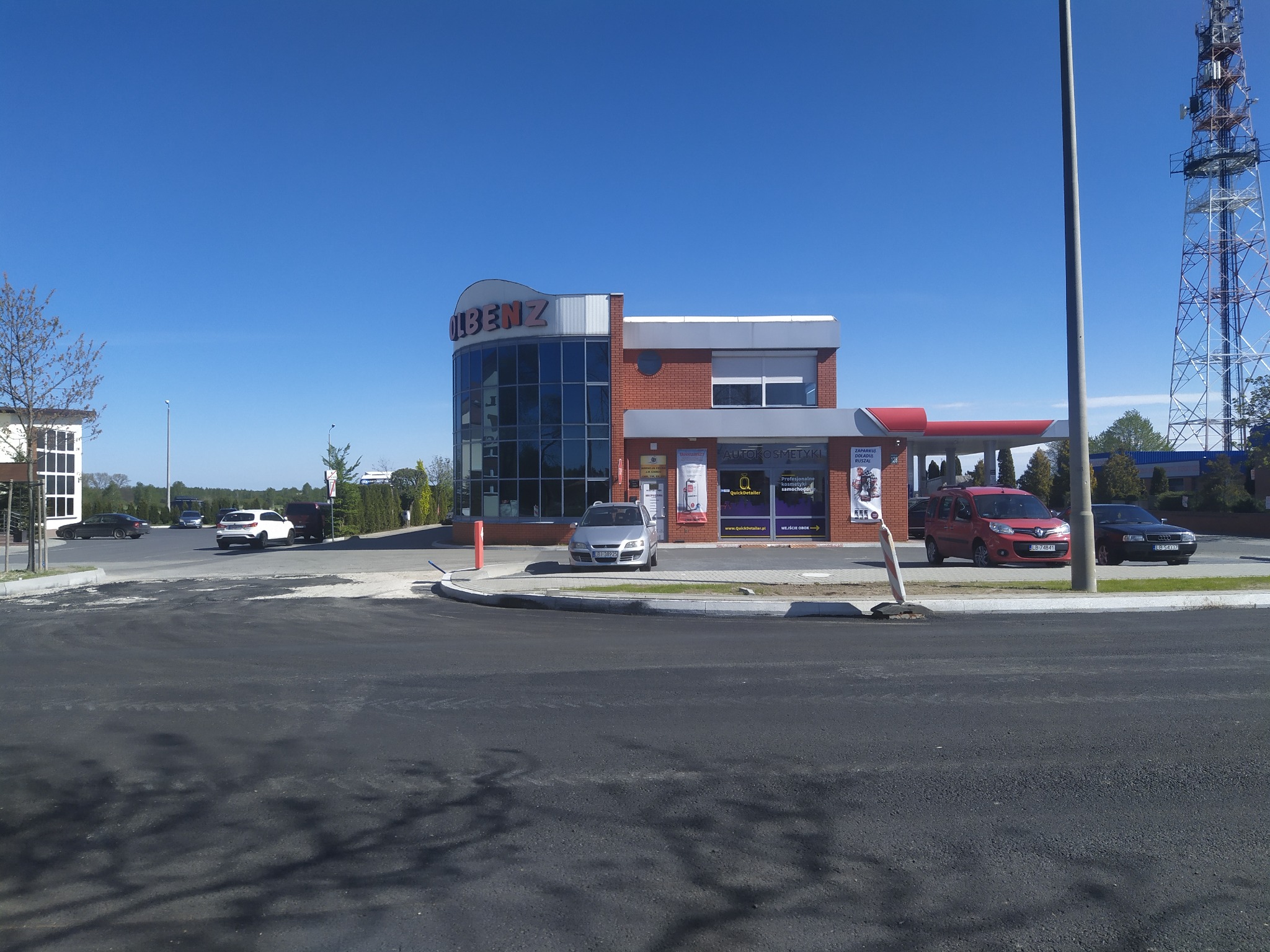 Stacja benzynowa z budynkiem usługowym Olbenz w słoneczny dzień, widoczne zaparkowane samochody i wieża telekomunikacyjna w tle.