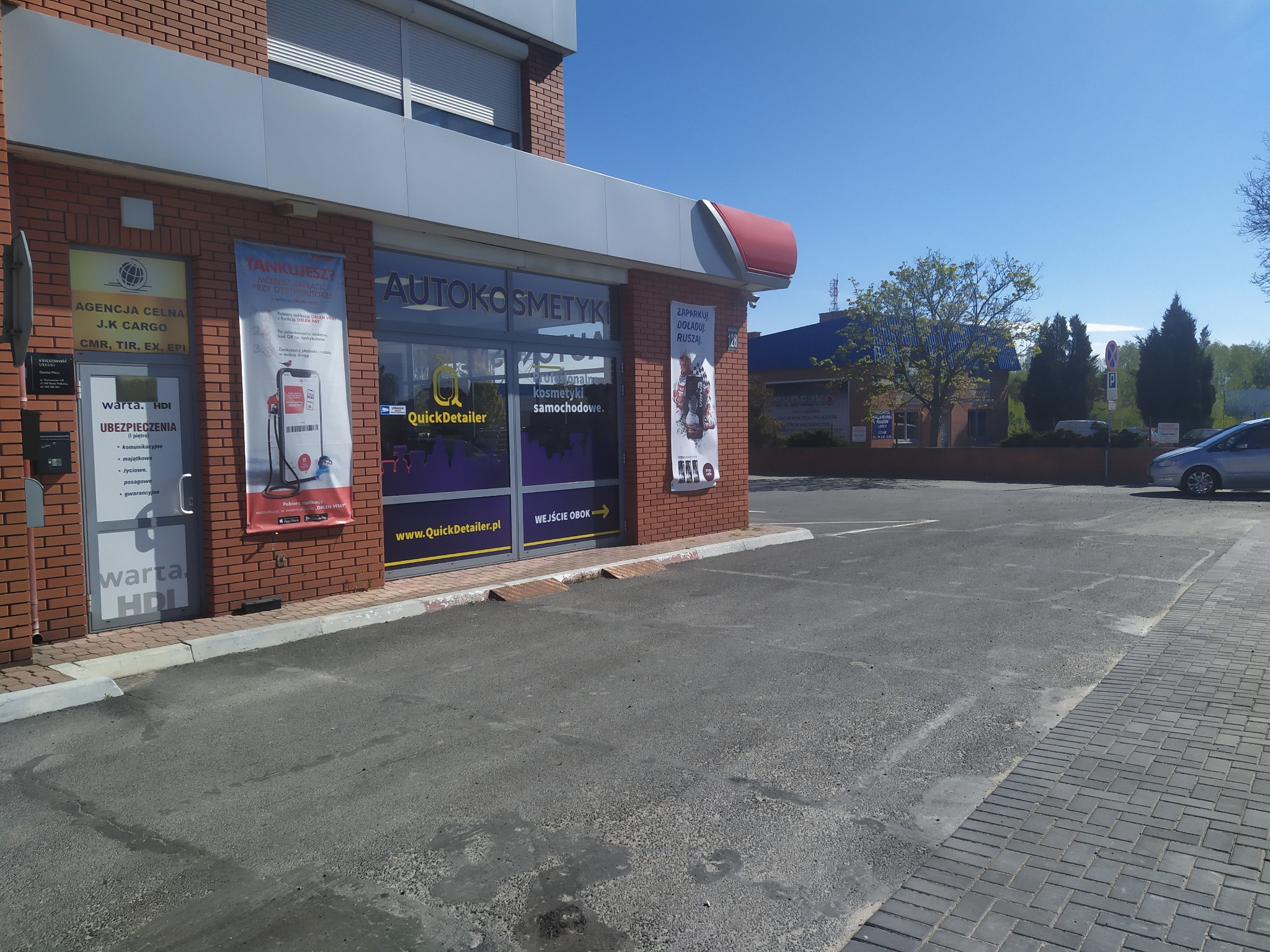 Budynek z szyldami Agencja Celna J.K Cargo, Autokosmetyki QuickDetailer oraz Warta HDI Ubezpieczenia, reklama Orlen Vitay na ścianie, parking przed budynkiem.