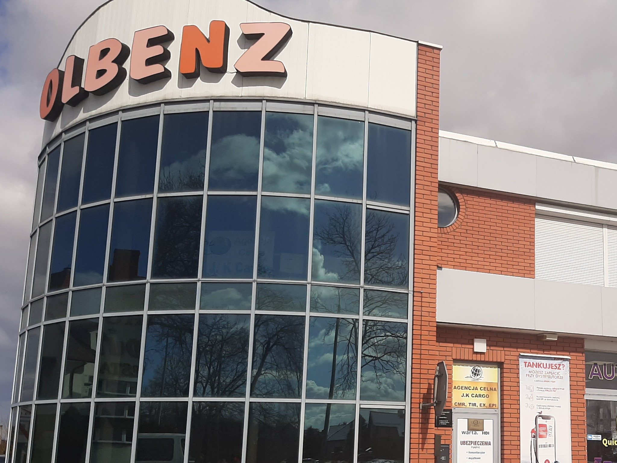 Budynek agencji celnej i ubezpieczeniowej Olbenz z dużymi oknami, w których odbija się błękitne niebo z chmurami. Na ścianie tabliczki informacyjne dotyczące agencji celnej J.K Cargo i Warta HDI...