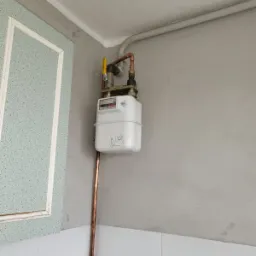 Gazomierz z miedzianymi rurami i żółtym zaworem, zamontowany na szarej ścianie z białą glazurą i fragmentem drzwi z dekoracyjną ramką.