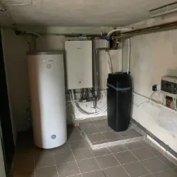 W kotłowni stoi biały zasobnik ciepłej wody marki LWAR, obok niego biały piec Panasonic, czarny zmiękczacz wody Carson oraz skrzynka elektryczna na ścianie z widocznymi kablami.