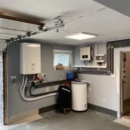 Instalacja urządzeń grzewczych w garażu: pracownik w czarnej koszulce Panasonic łączy biały zbiornik ciepłej wody użytkowej, obok zawieszony na ścianie moduł i inwerter. Widoczne rury instalacyjne,...