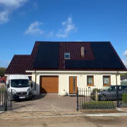 Dom jednorodzinny z ciemnobrązową dachówką i panelami słonecznymi na dachu, biała elewacja, brama wjazdowa, samochód osobowy i dostawczy na posesji, błękitne niebo.