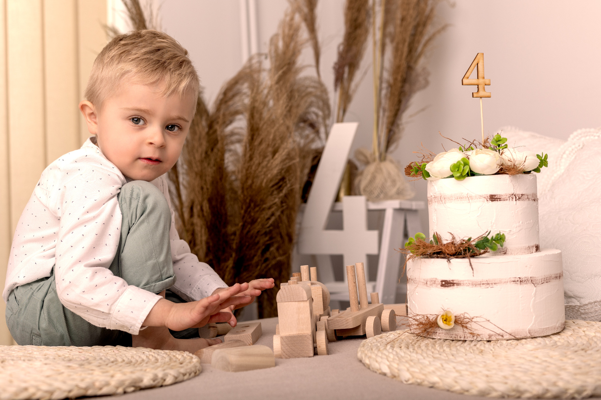 Uroczy chłopiec w jasnej koszuli i miętowych spodniach bawi się drewnianym pociągiem obok dwupiętrowego tortu z numerem 4, całość w naturalnej kolorystyce z elementami dekoracyjnymi z suszonych traw.