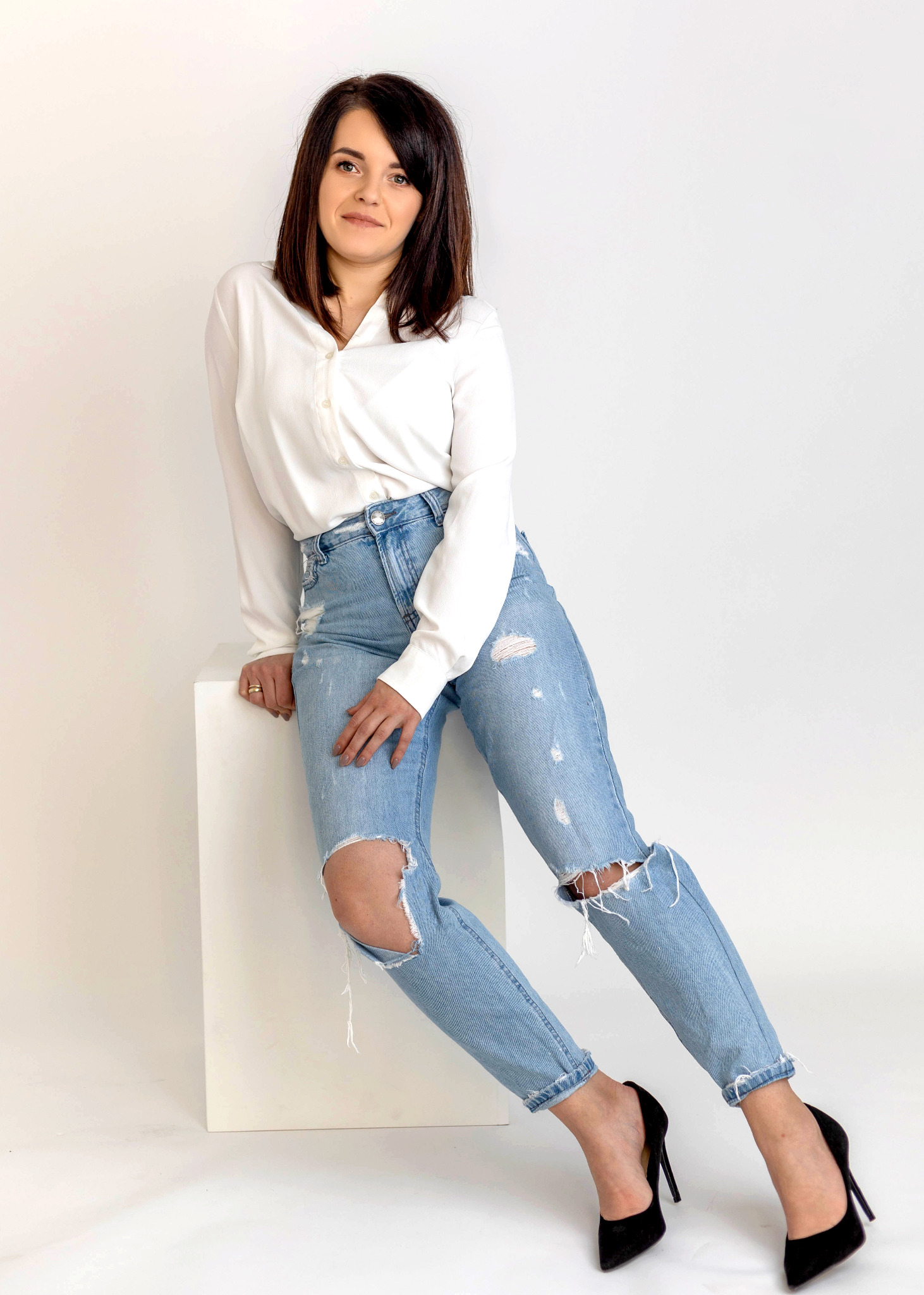 Portret kobiety w białej koszuli i podartych jeansach, opierającej się o biały sześcian, na białym tle.