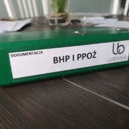 Zielony segregator z napisem 'Dokumentacja BHP i PPOŻ' oraz logo firmy Lider BHP, leżący na drewnianym stole, w tle widoczna roślina w szklanym wazonie.