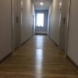 Długi, pusty korytarz biurowy z drewnopodobną podłogą, białymi ścianami i rzędem drzwi po obu stronach, oświetlony światłem z okna na końcu korytarza i wbudowanymi lampami sufitowymi.