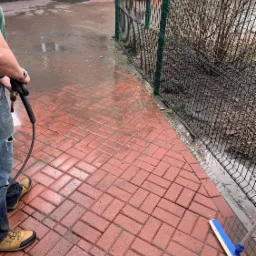 Czerwona kostka brukowa czyszczona myjką ciśnieniową, widoczne mokre plamy i osoba w jeansach i brązowych butach z dyszą myjącą, obok zielone ogrodzenie i budynek.