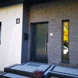 Nowoczesne wejście do domu z grafitową elewacją, ciemnymi oknami i drzwiami, widoczne schody z kostki brukowej i numer domu '4'. W szybach okien odbija się jesienne drzewo i zaparkowany samochód.