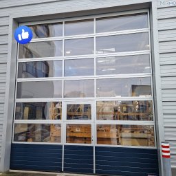 MIRMO sp. z o.o. - Brama garażowa segmentowa z przeszkleniami, widoczna z zewnątrz. Dolny panel w kolorze granatowym, reszta w srebrnej ramie. W tle widoczny podnośnik.