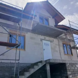 Dom w trakcie budowy z oknami w drewnianych ramach, widoczne rusztowania i białe drzwi z logo. Słońce w tle, błękitne niebo.