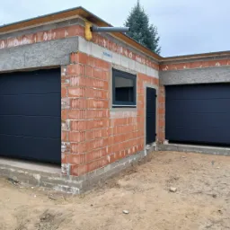 Garaż z dwiema ciemnymi bramami segmentowymi w surowym stanie, z widoczną konstrukcją z cegieł i betonowym nadprożem. Widoczne okno i drzwi wejściowe.