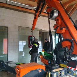 MIRMO sp. z o.o. - Montaż okien z użyciem specjalistycznego dźwigu przyssawkowego. Dwóch monterów nadzoruje proces, widoczne surowe mury budynku w trakcie budowy.