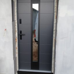 MIRMO sp. z o.o. - Nowoczesne, szare drzwi zewnętrzne z przeszkleniem, zamontowane w surowym otworze. Widoczne uszczelnienia i fragment białego busa w odbiciu szyby.