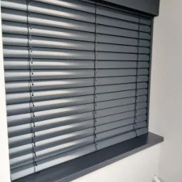 Szare żaluzje poziome, zamontowane w oknie z szarą ramą, widoczne z bliska, z delikatnym światłem padającym na lamele.