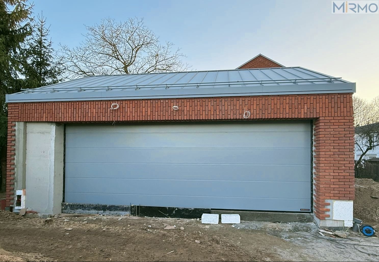 Szara brama garażowa segmentowa Hörmann z panelami micrograin, zamontowana w garażu z cegły klinkierowej pod szarym dachem. Widoczne kable instalacyjne na ścianie.