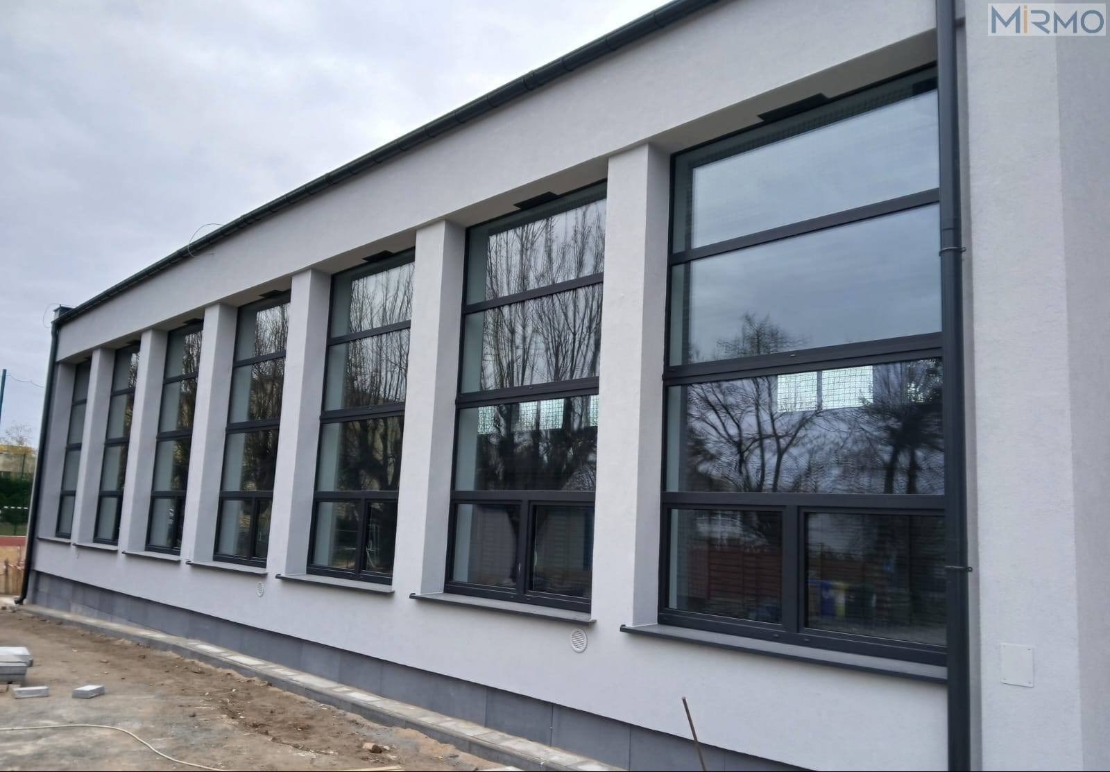 Elewacja budynku z rzędem dużych, czarnych okien aluminiowych, odbijających drzewa i niebo. Widoczne detale architektoniczne i częściowo przygotowany teren.