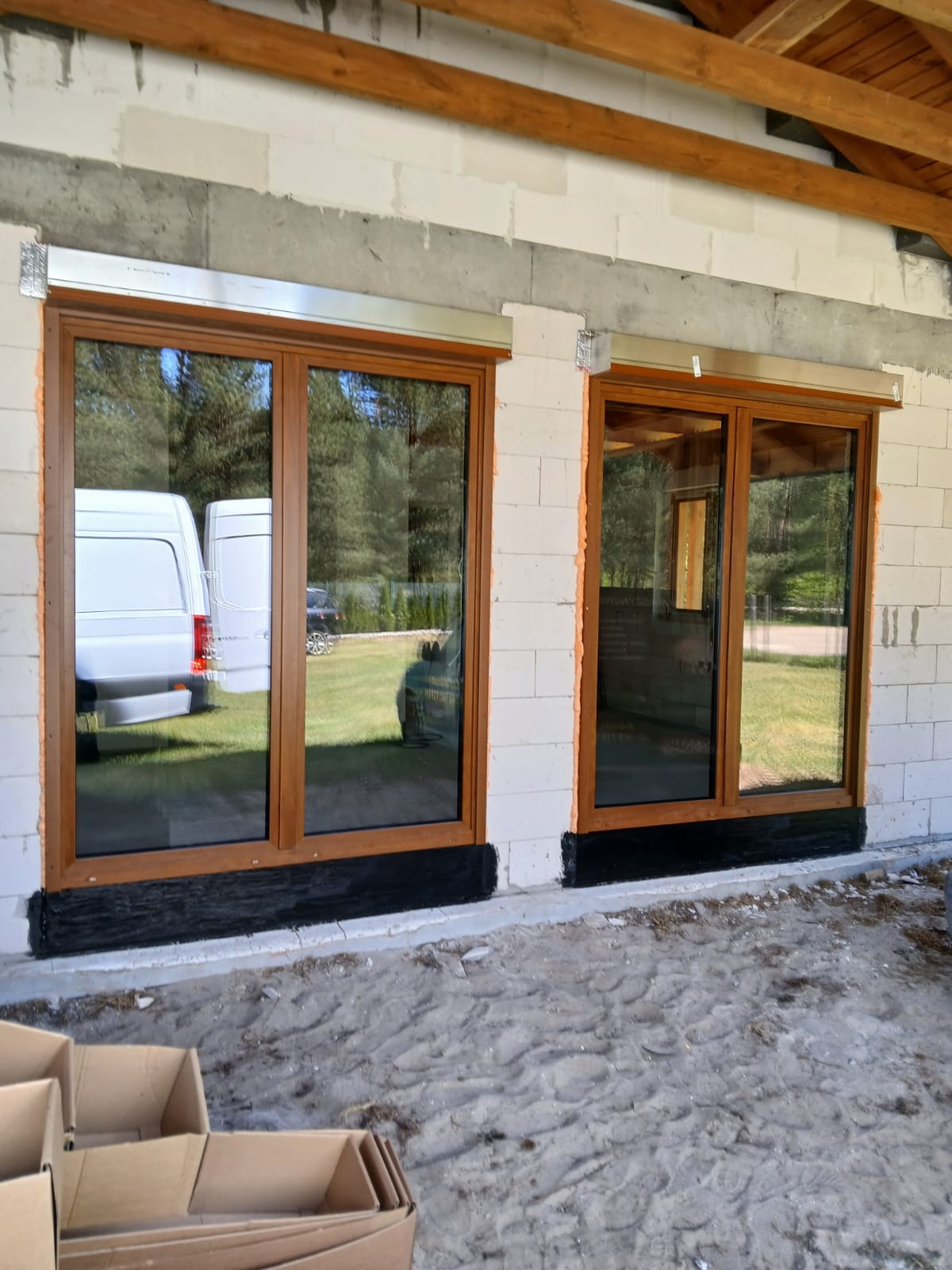 Dwa brązowe okna z roletami zewnętrznymi w surowym murze z betonu komórkowego. Widoczne zabezpieczenie przeciwwilgociowe u dołu i fragmenty pianki montażowej.