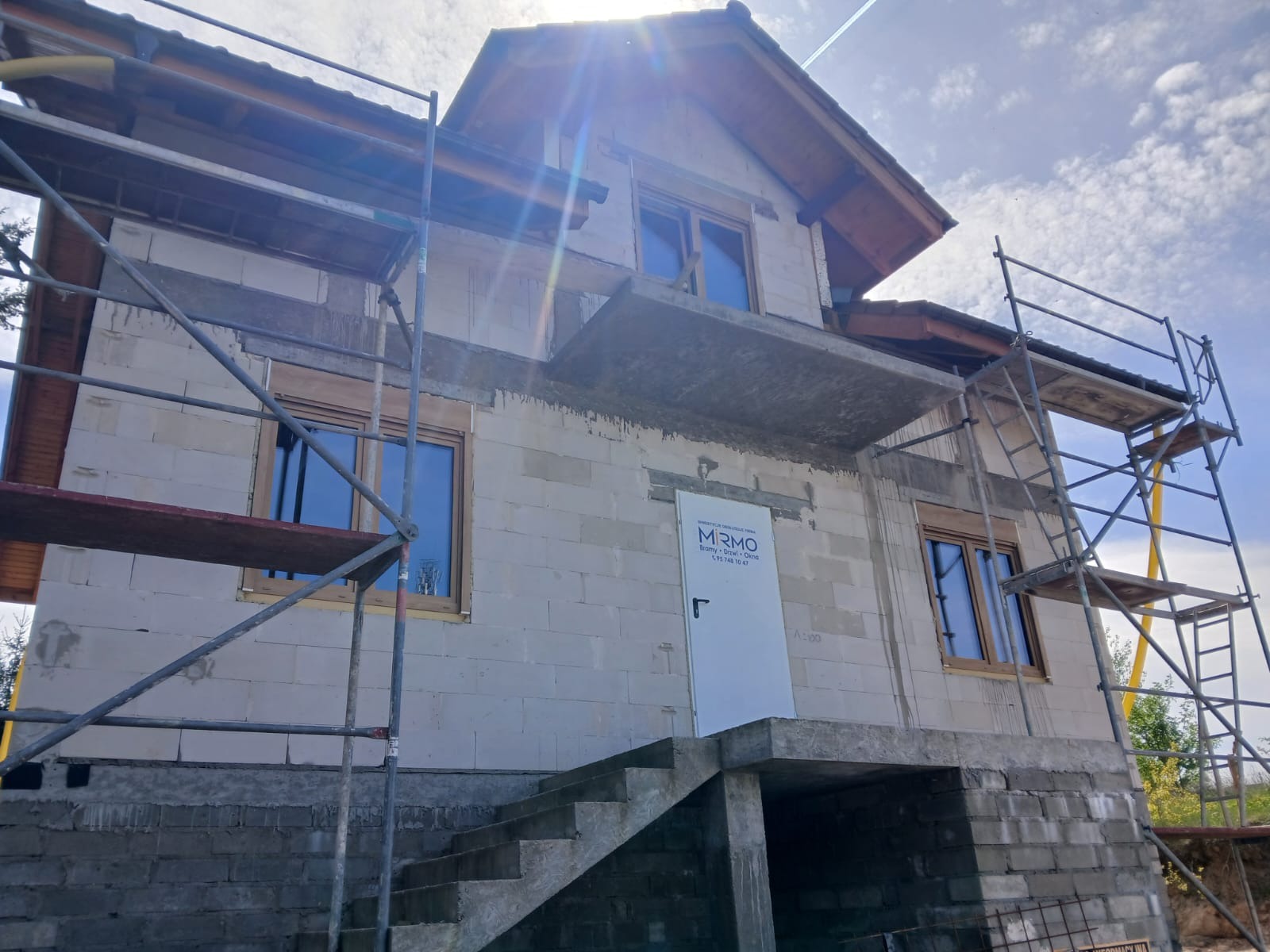 Dom w trakcie budowy z oknami w drewnianych ramach, widoczne rusztowania i białe drzwi z logo. Słońce w tle, błękitne niebo.