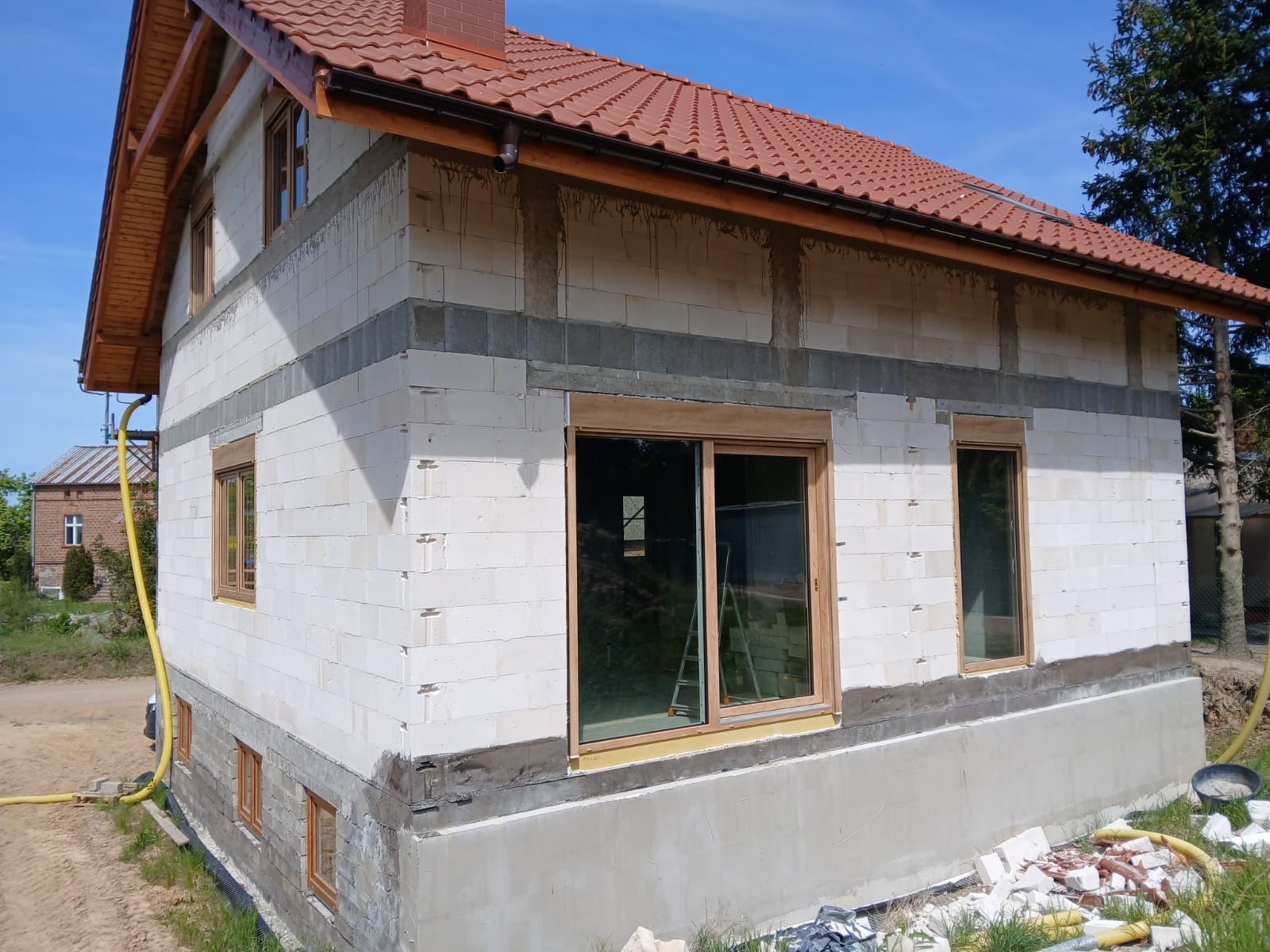 Budowa domu z białego bloczka, widoczne wstawione okna z drewnianą ramą i czerwony dach. Na pierwszym planie gruz budowlany. Słoneczny dzień.