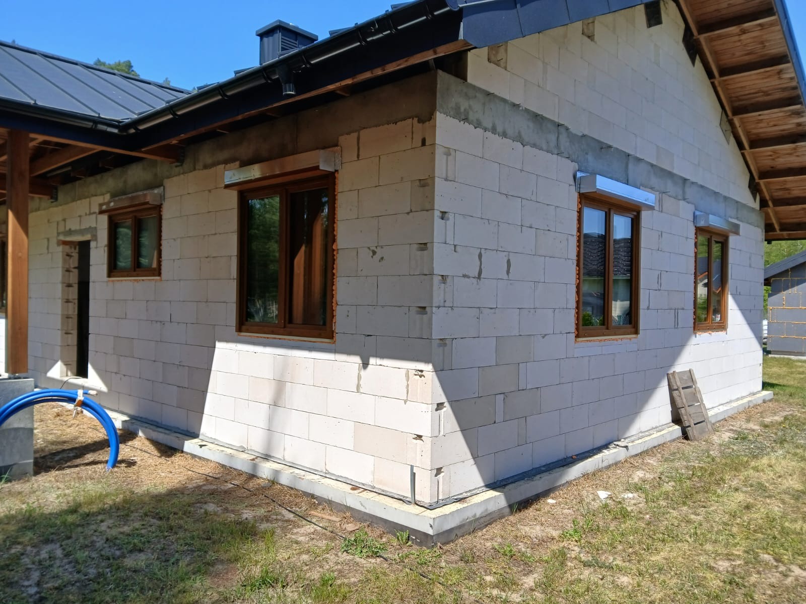 Surowy stan budynku z oknami z brązowymi ramami, widoczne bloczki betonowe, fragment dachu i trawnik. Budowa domu w trakcie realizacji.