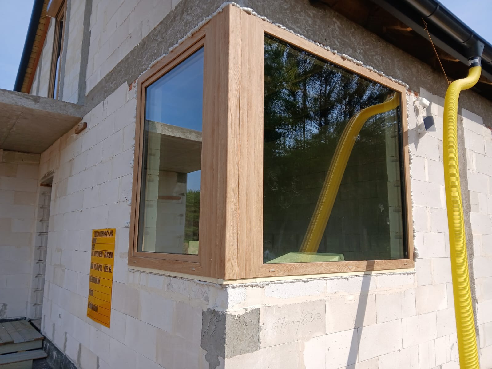Narożne okno z drewnianą ramą w surowym budynku. Widoczna żółta rura wentylacyjna i tablica informacyjna. Okno od zewnątrz, widok na budowę.