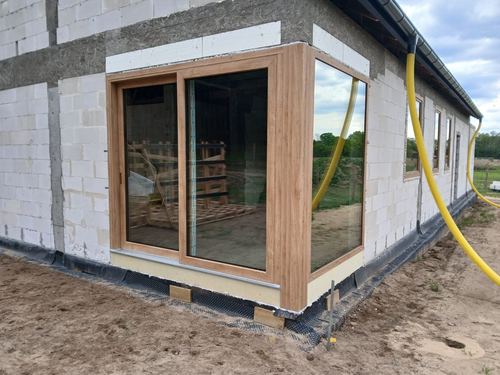 Zamontowane okno przesuwne z drewnianą ramą w surowym budynku z białego bloczka. Widoczny fragment elewacji z orynnowaniem i żółtym wężem odprowadzającym wodę.