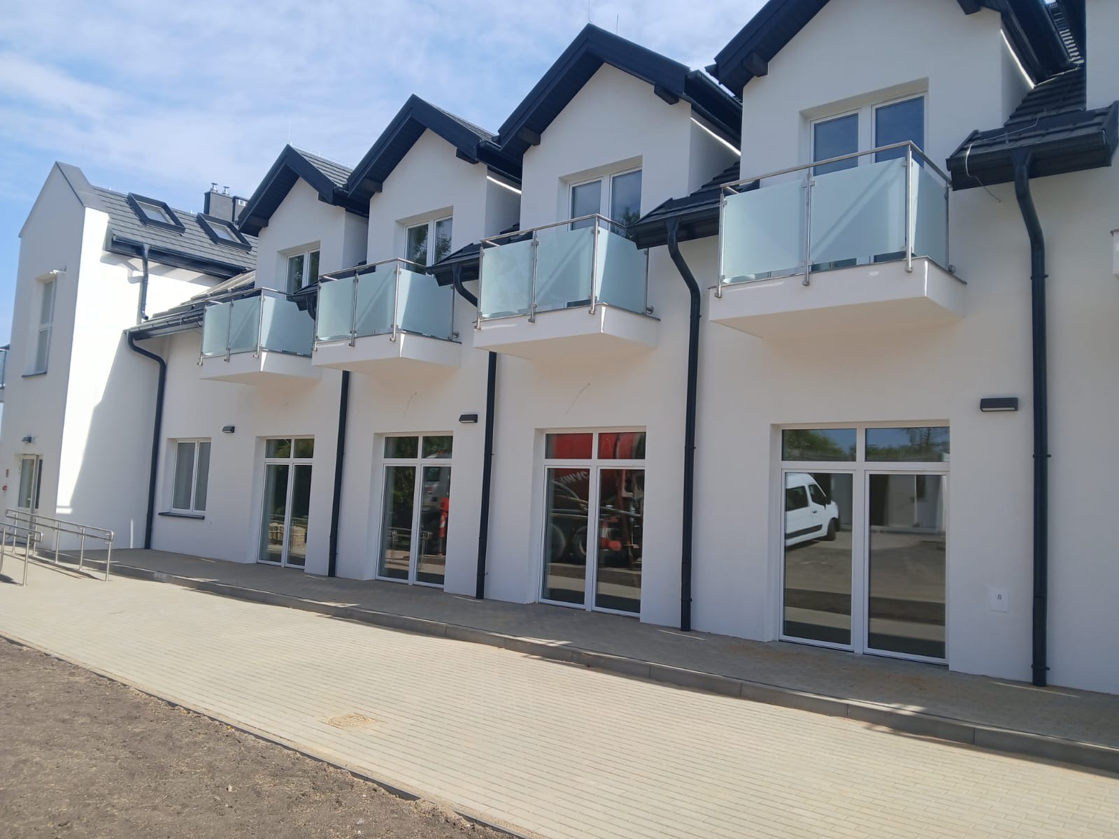 Biały budynek z balkonami i szklanymi balustradami. Widoczne okna i drzwi z białymi ramami. Jasne, słoneczne oświetlenie.