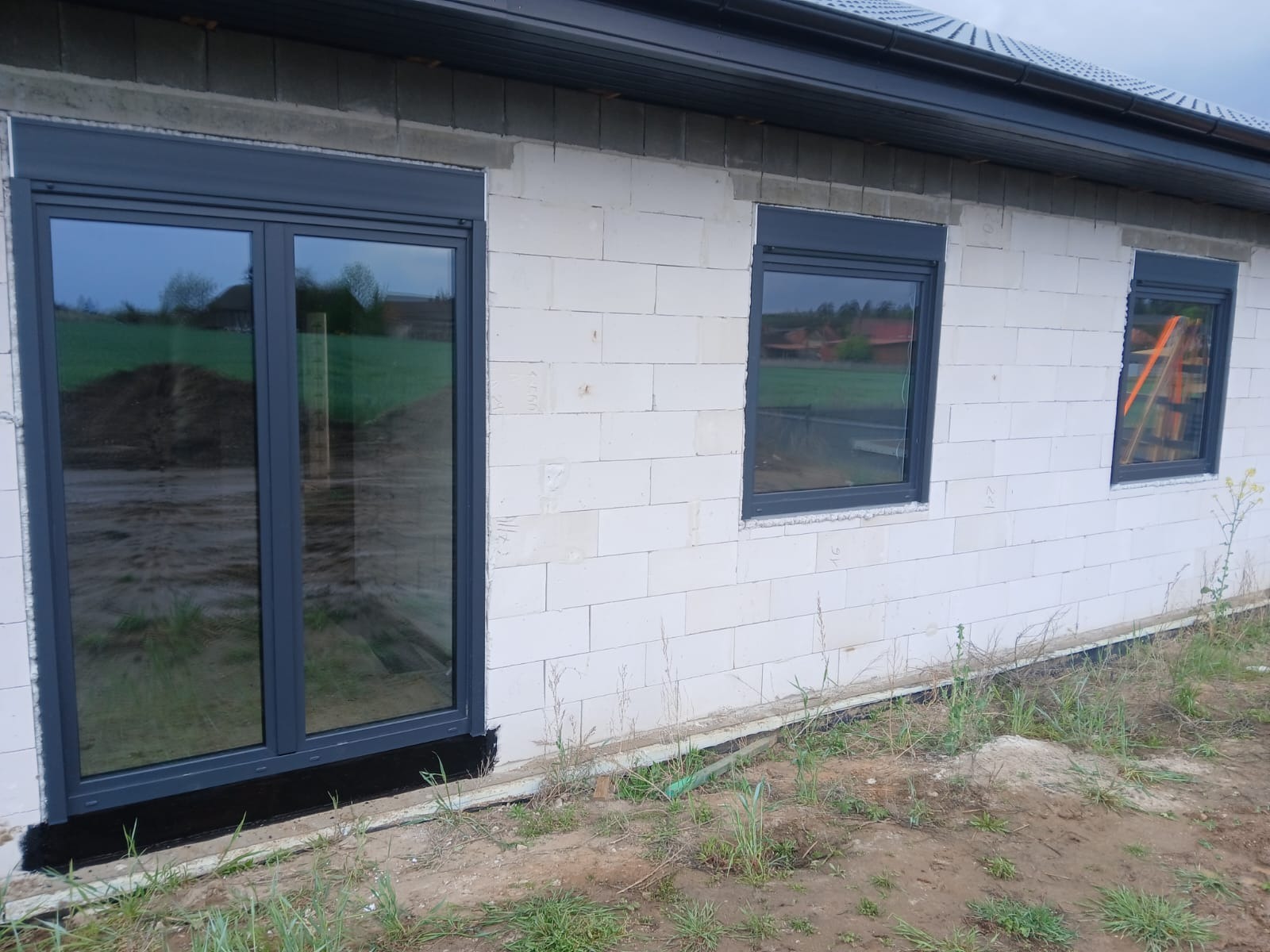 Szare okna PCV z roletami zewnętrznymi w surowym budynku z białego bloczka. Widoczne drabiny w odbiciu szyby. Okna zamontowane na czarnym pasie hydroizolacji.