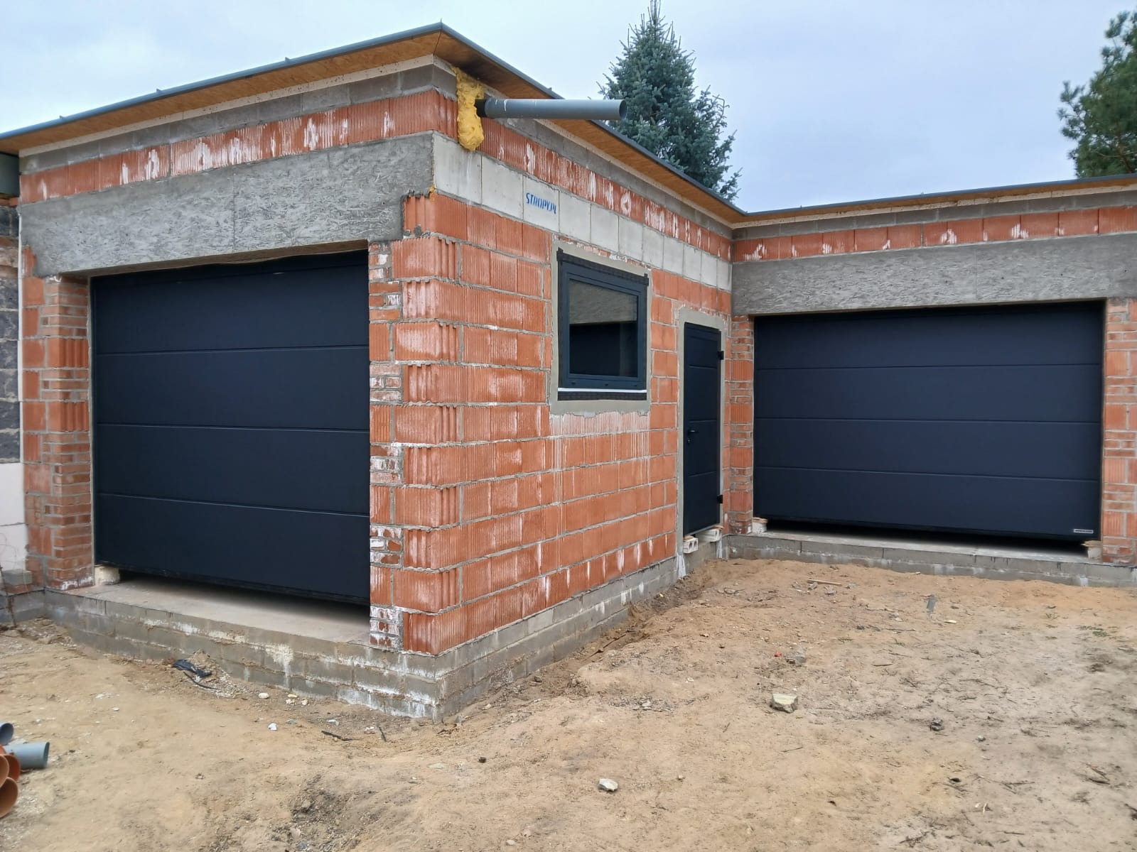 Garaż z dwiema ciemnymi bramami segmentowymi w surowym stanie, z widoczną konstrukcją z cegieł i betonowym nadprożem. Widoczne okno i drzwi wejściowe.