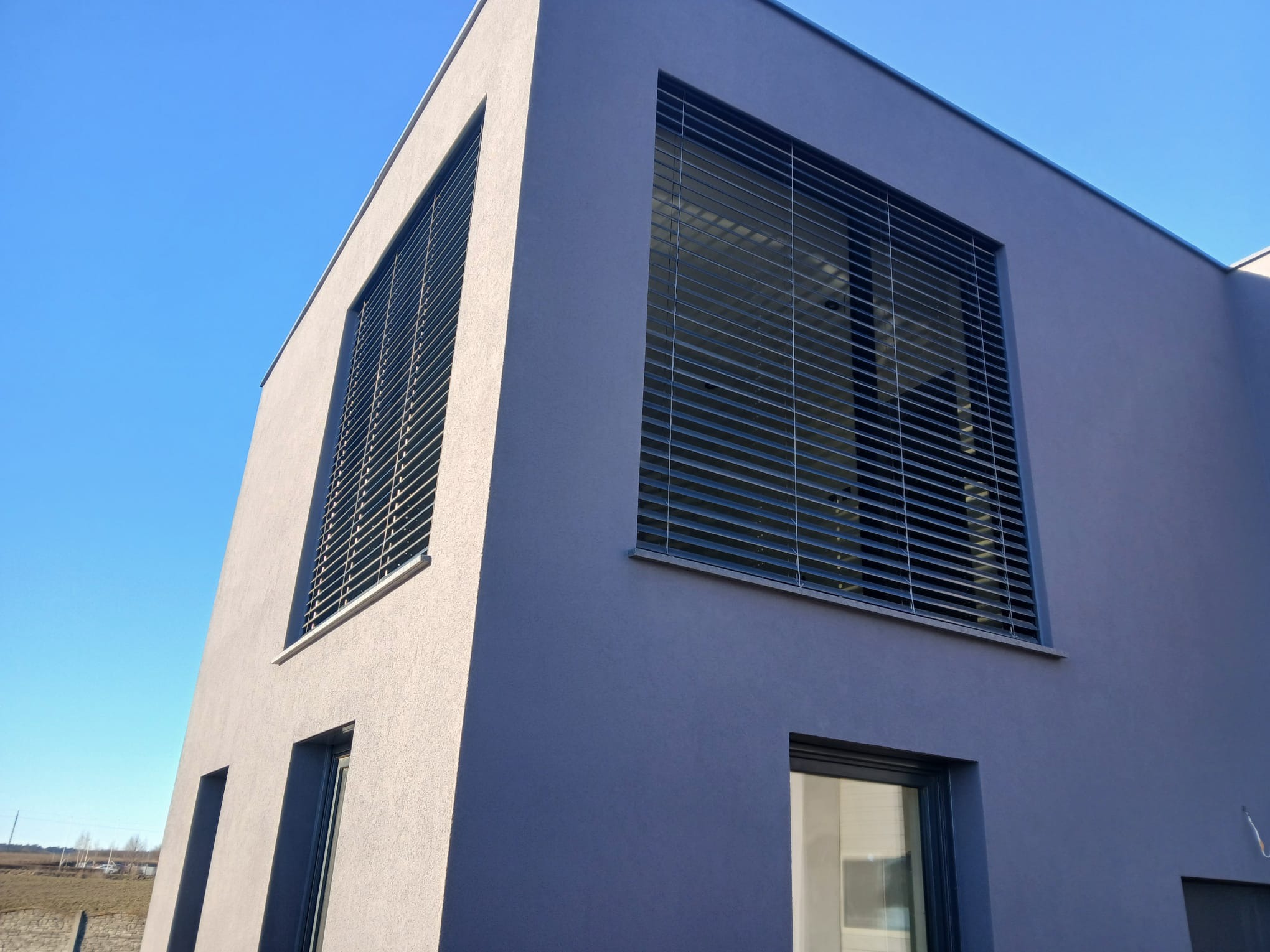 Szare rolety zewnętrzne na oknach nowoczesnego domu w słoneczny dzień. Ujęcie z dołu, uwydatniające architekturę budynku i detale rolet.