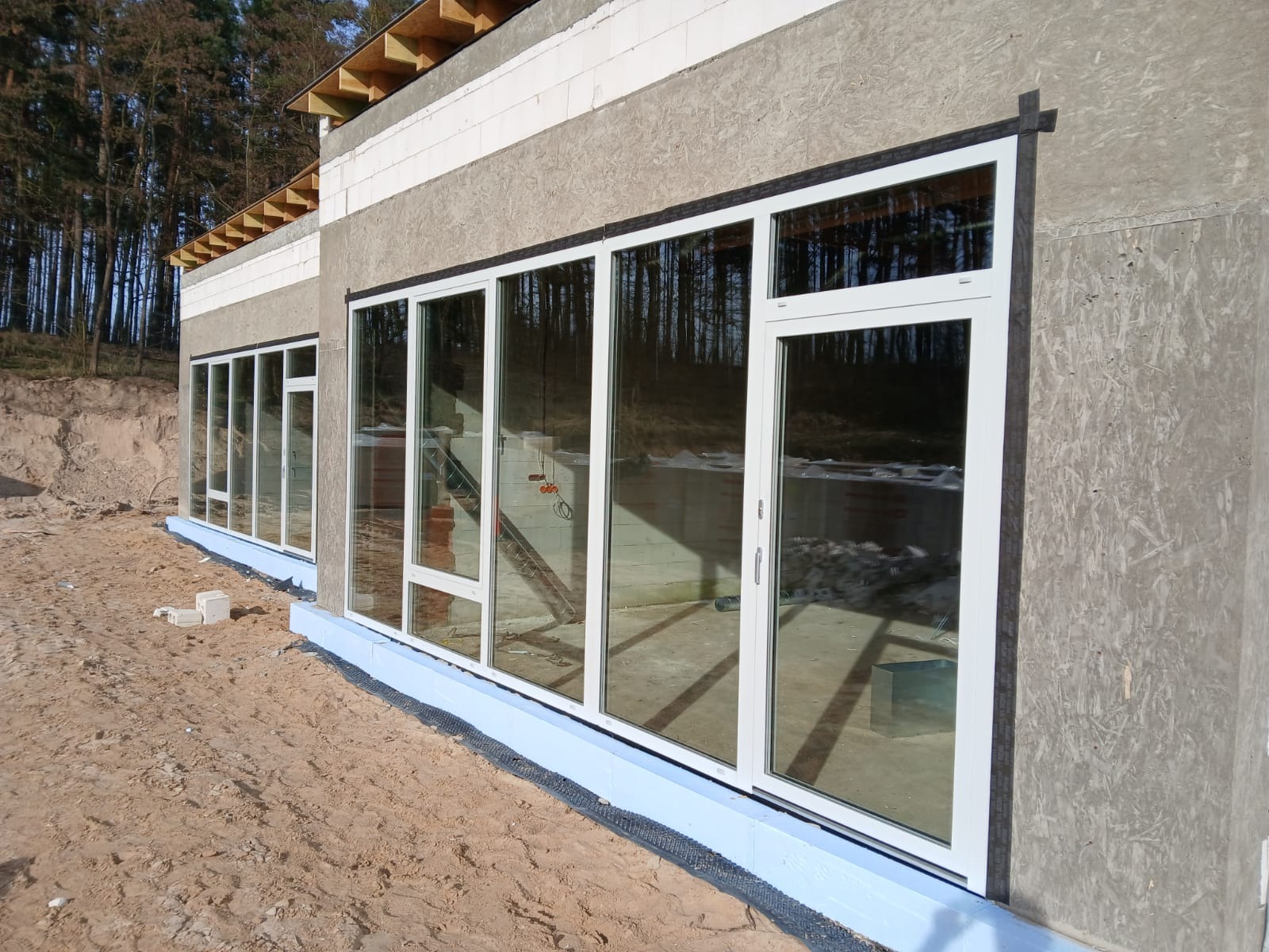 Nowoczesne okna aluminiowe z białymi ramami w surowym stanie budynku. Widoczne ocieplenie fundamentów i las w odbiciu szyb. Jasne, słoneczne oświetlenie.