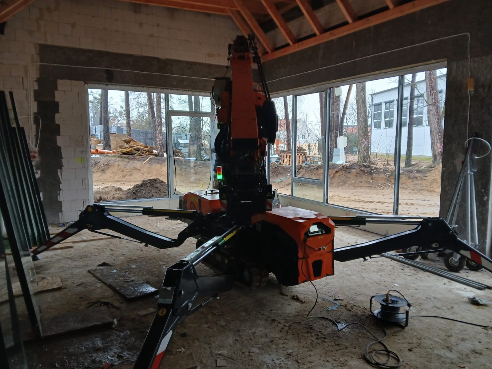 Pomarańczowy mini żuraw pająk w surowym wnętrzu budynku z dużymi oknami, widoczne elementy konstrukcyjne dachu. W tle drzewa i budynki.