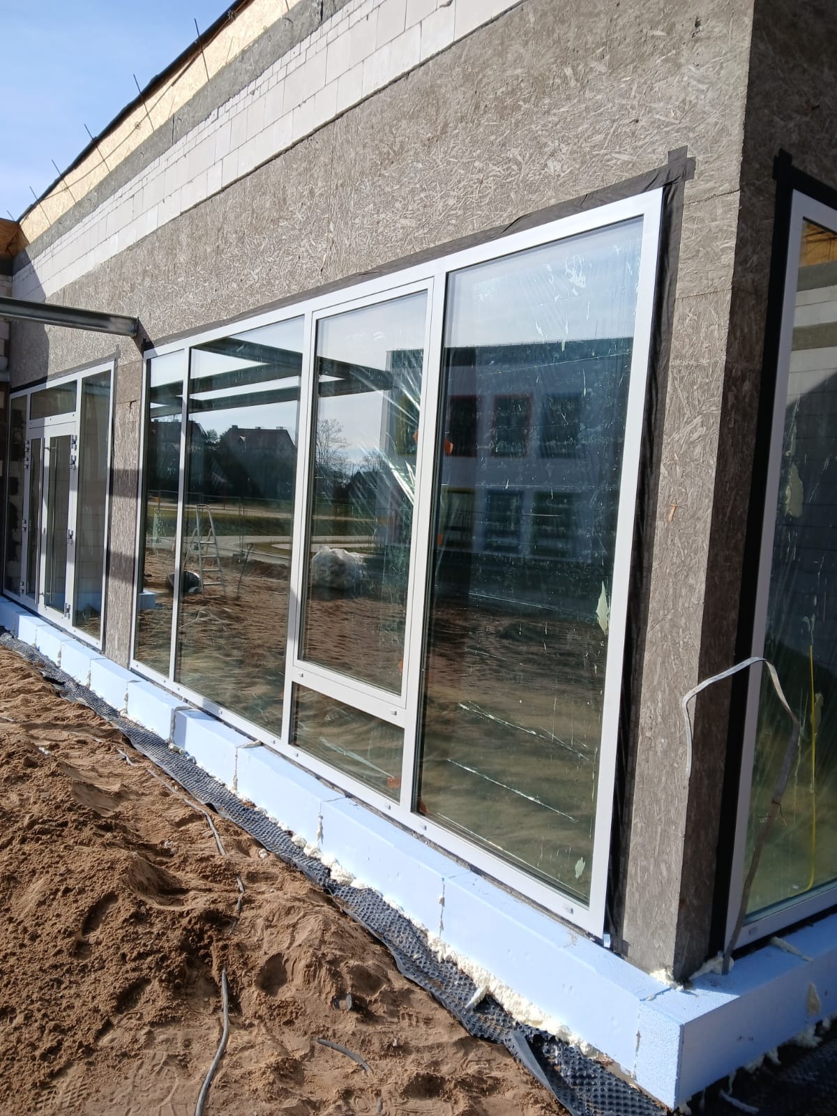 Montaż okien aluminiowych z białymi ramami w surowym stanie budynku, widoczne z zewnątrz w Międzychodzie. Część okien zabezpieczona folią.