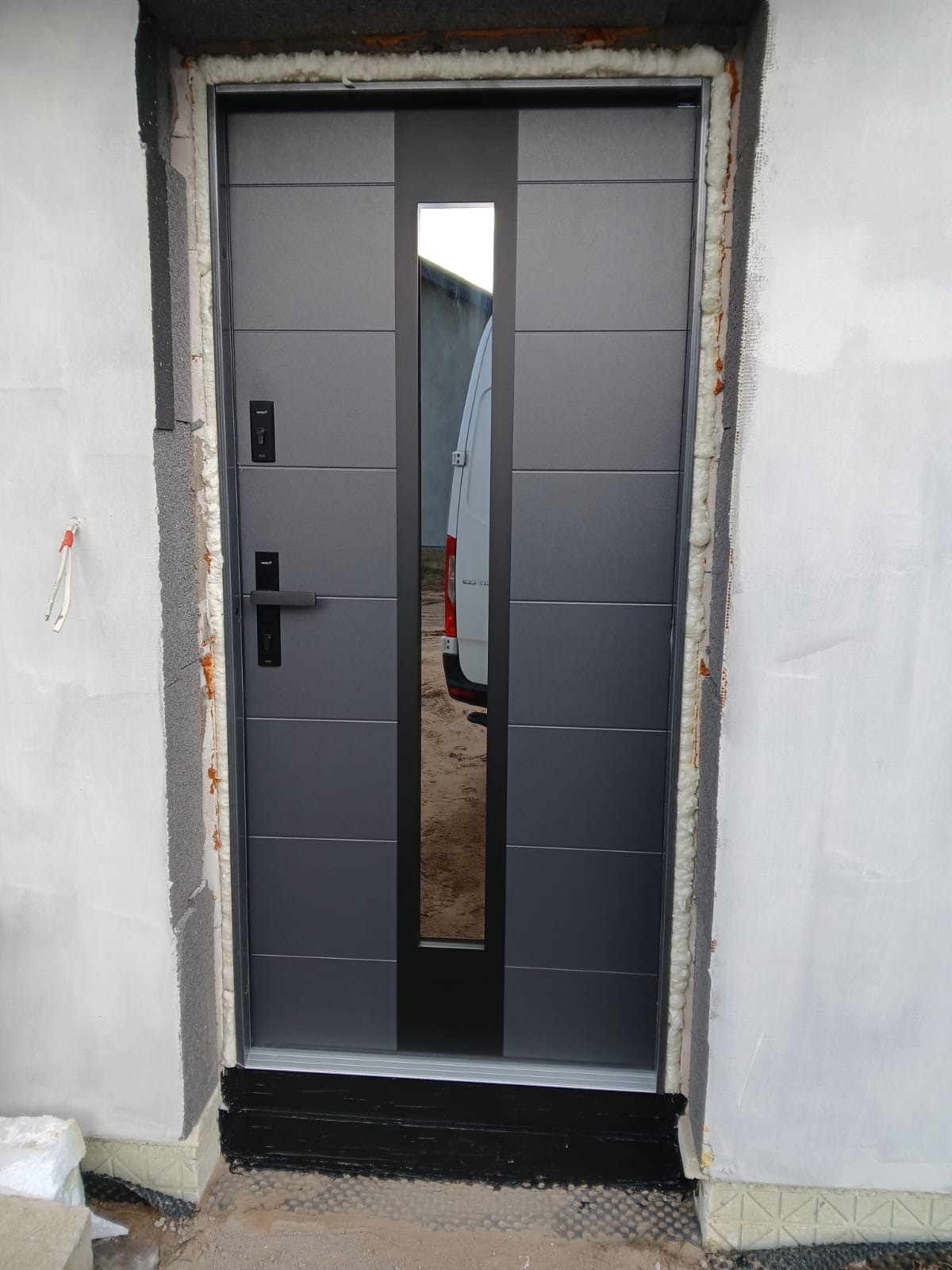Nowoczesne, szare drzwi zewnętrzne z przeszkleniem, zamontowane w surowym otworze. Widoczne uszczelnienia i fragment białego busa w odbiciu szyby.