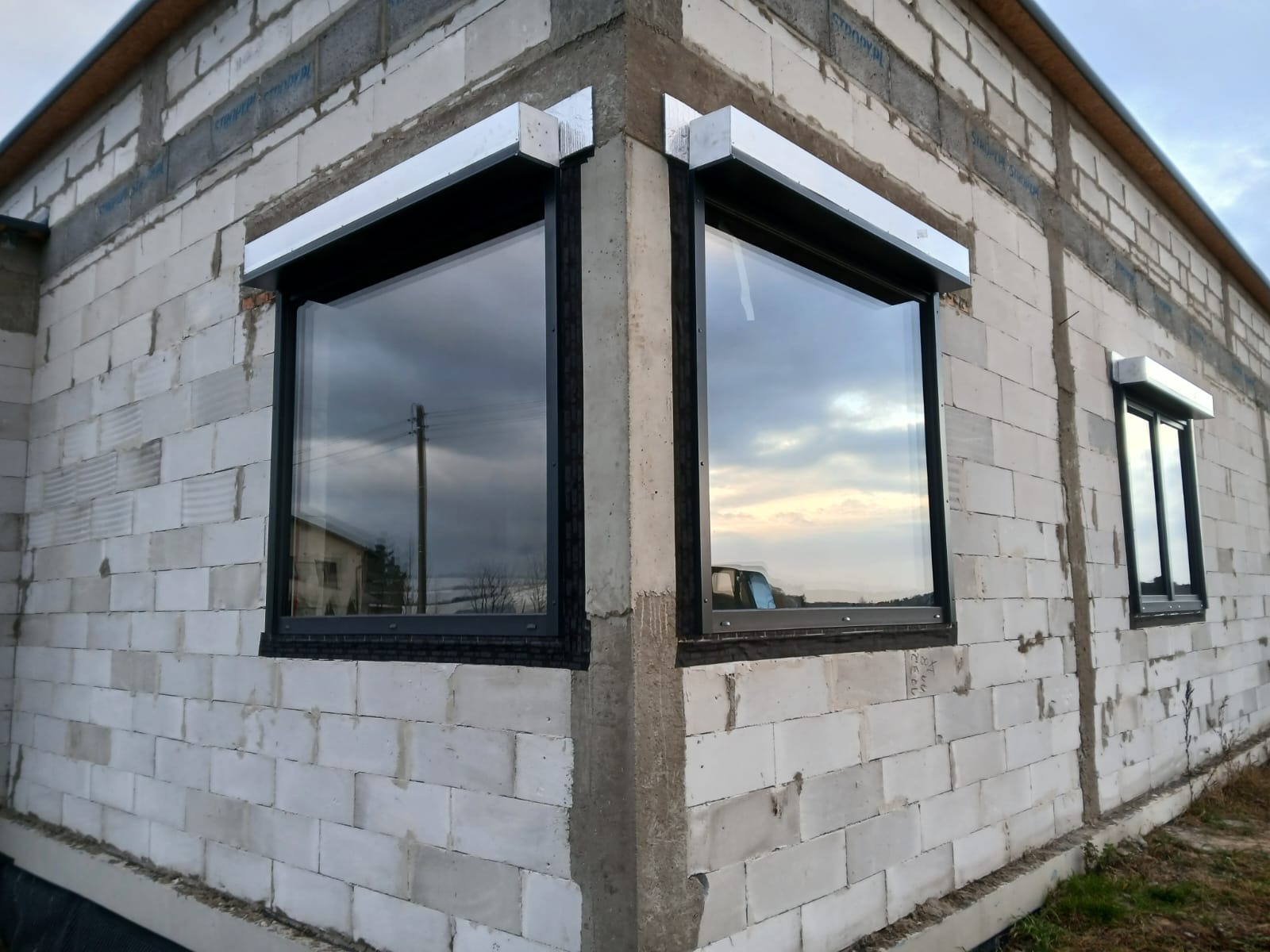 Czarne ramy okienne z roletami zewnętrznymi w surowym budynku z białych bloczków. Widok z zewnątrz na dwa okna narożne i jedno mniejsze okno z boku.