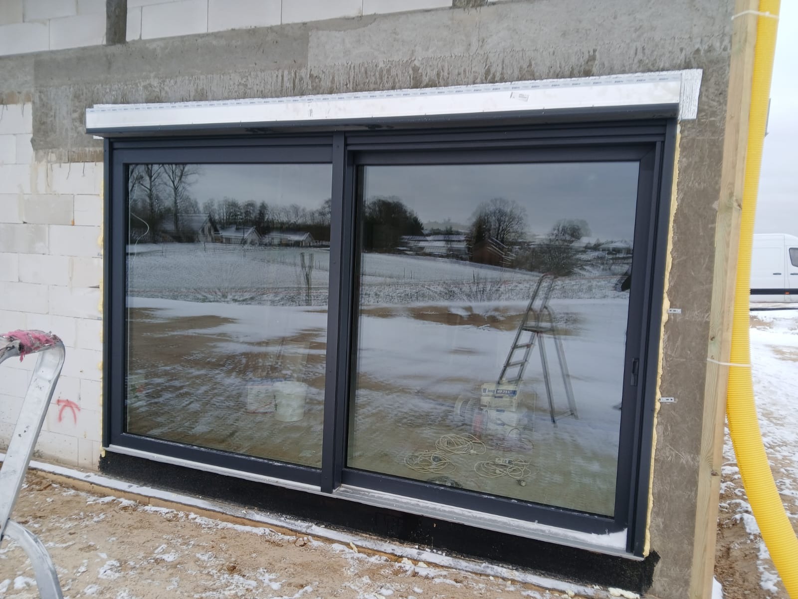 Duże, przesuwne okno w ciemnej ramie, zamontowane w surowym murze. Widoczny fragment rolety zewnętrznej. Na zewnątrz zimowy krajobraz i drabina oparta o okno.