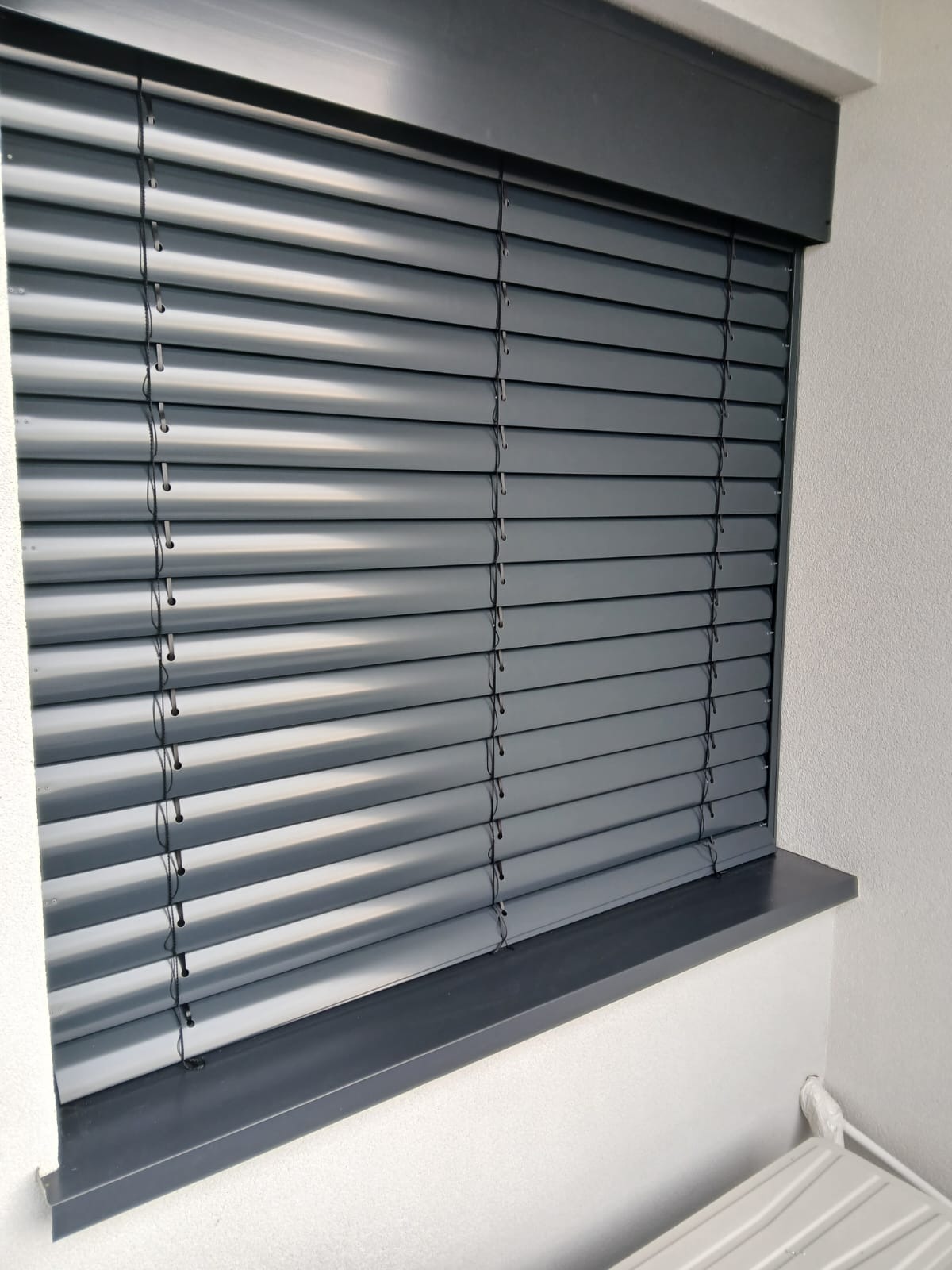 Szare żaluzje poziome, zamontowane w oknie z szarą ramą, widoczne z bliska, z delikatnym światłem padającym na lamele.