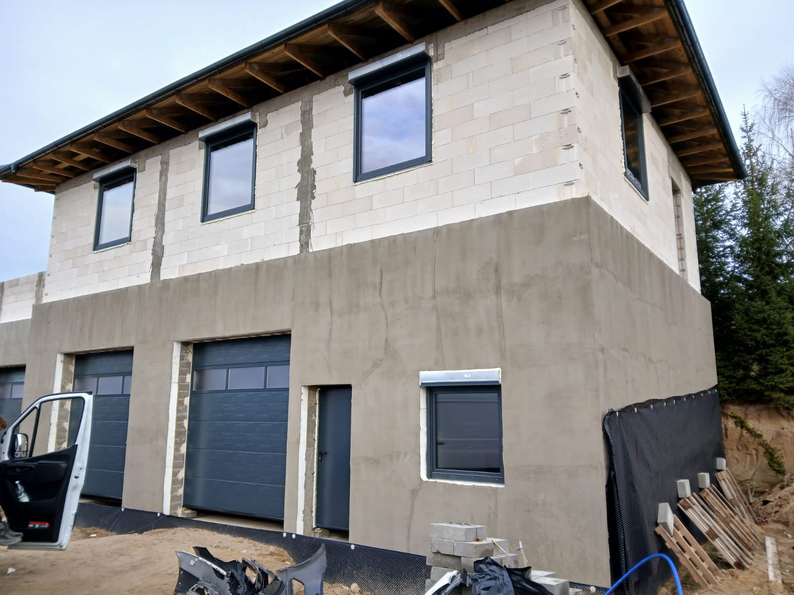 Nowy budynek w trakcie wykończenia: szare bramy garażowe, okna z roletami zewnętrznymi, otynkowana część elewacji, widoczne bloczki z betonu komórkowego.